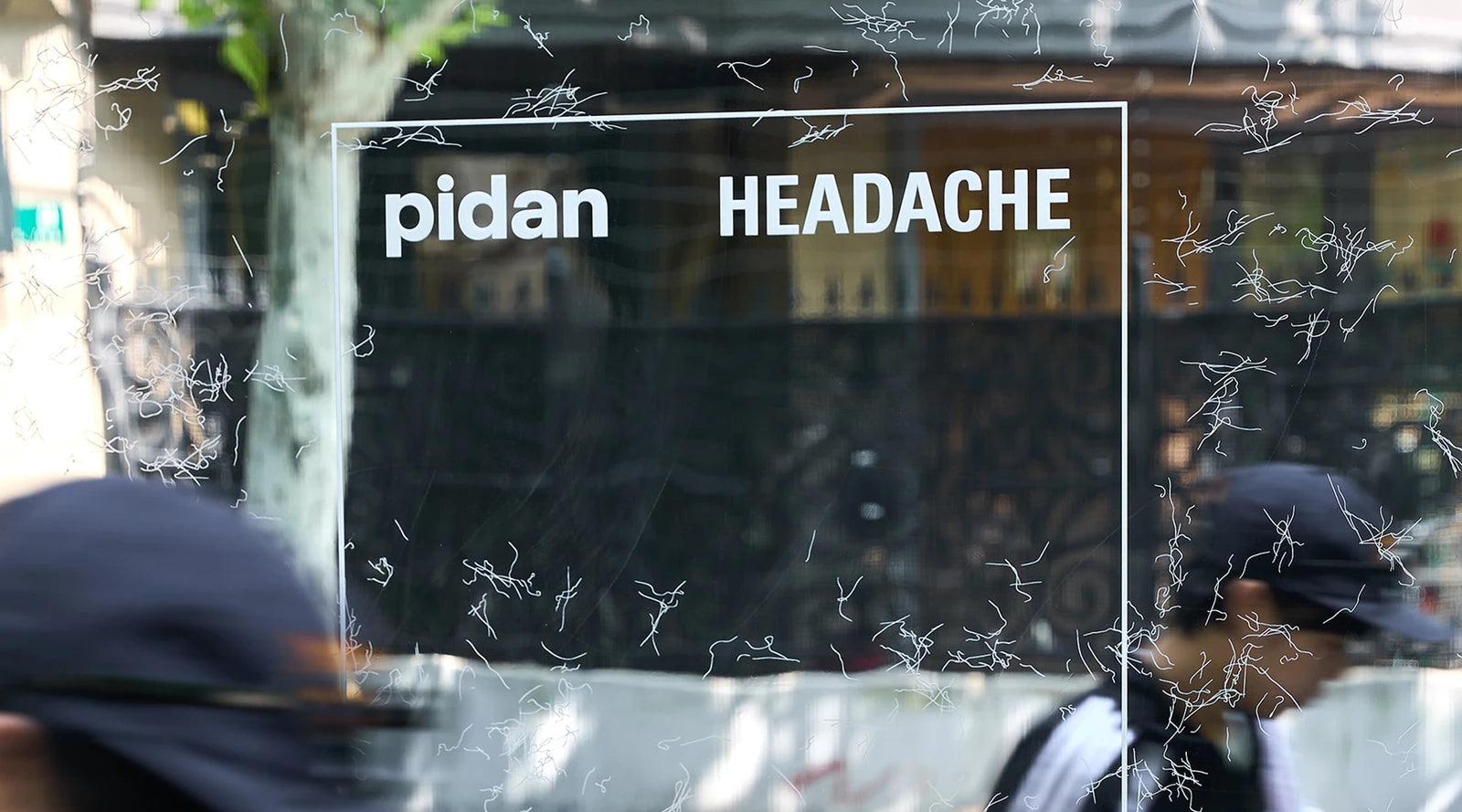 A pop-up shopfront window carrying the pidan and HEADACHE lockup, hand-drawn strands printed across the glass, passers-by blurred in front